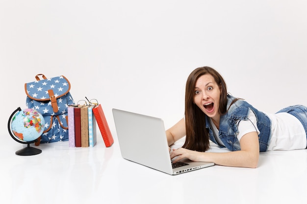 Young shocked woman student in denim clothes working on laptop pc computer lying near globe, backpack, school books isolated