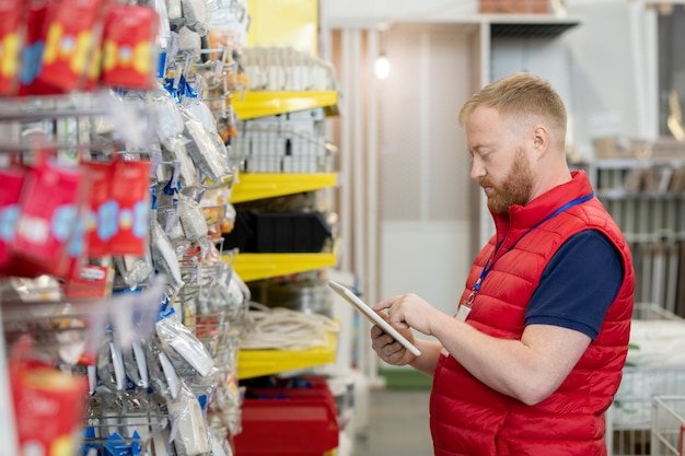 Young shop assistant in red uniform using tablet during work in warehouse