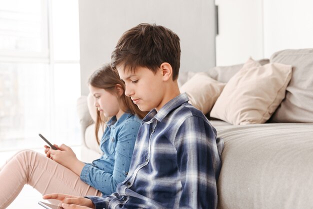 Young siblings girl and boy sitting on floor near sofa at home, and both using smartphone