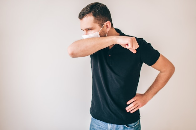 Giovane uomo malato isolato sopra la parete. persona di sesso maschile in camicia nera che tossisce attraverso la mascherina di protezione medica. coprire la bocca con la mano.