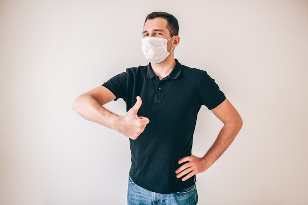 Young sick man isolated over wall. Positive guy in black shirt hold big thumb up. Young man wear white protection medical mask to avoid sickness.