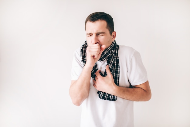 Photo young sick man isolated over white wall. guy with scarf around neck. coughing out loud. hold fist close to mouth. suffer from ache and chest pain.
