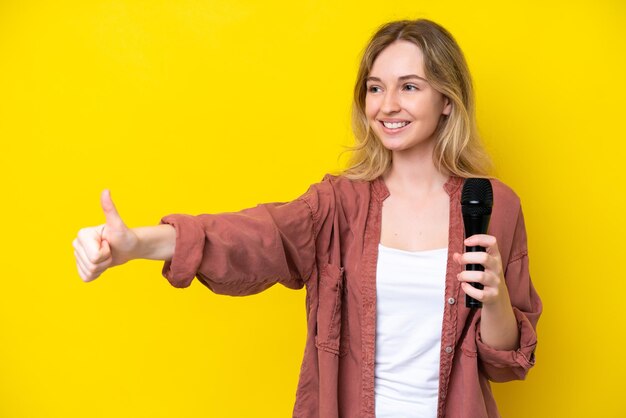 Young singer caucasian woman picking up a microphone isolated on yellow background giving a thumbs up gesture