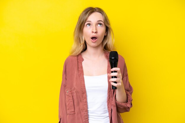 Young singer caucasian woman picking up a microphone isolated on yellow background looking up and with surprised expression