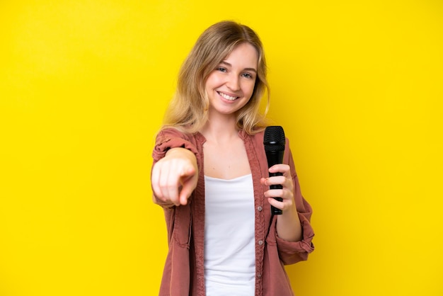 Giovane cantante donna caucasica che prende un microfono isolato su sfondo giallo che punta davanti con felice espressione