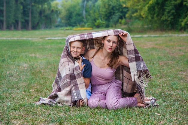 Young sister and brother with freckles on their faces stand covered with plaid in park, get warm, smiling and looking at camera.