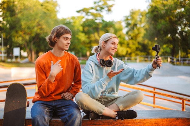 Ragazzo giovane skater in pullover arancione e ragazza con le cuffie che registrano un nuovo video per vlog mentre trascorrono felicemente del tempo insieme allo skatepark