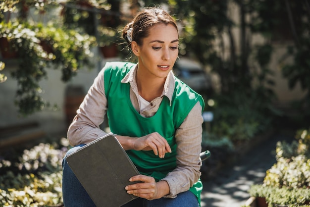 Young skilled woman holding digital tablet and checking of potted plants in a greenhouse.