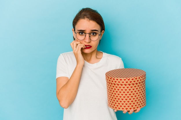 Young skinny arab girl holding a valentines day box biting fingernails, nervous and very anxious.