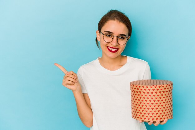 Young skinny arab girl holding a valentines day box smiling and pointing aside, showing something at blank space.