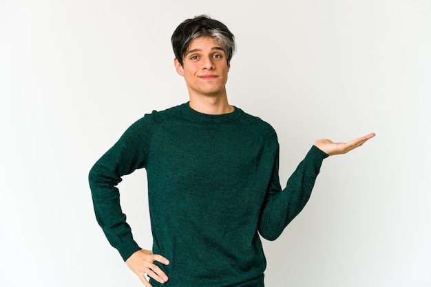 Young skinny hispanic man showing a copy space on a palm and holding another hand on waist.