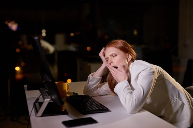 Young sleepy caucasian female at work place in office at night, need more rest, has no more strength to work at late night. side view on exhausted woman in formal wear yawning, tired of work