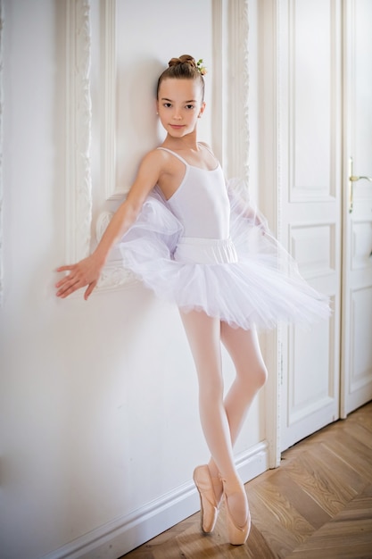 Young slender ballerina in a white tutu dances on pointe shoes in a spacious, bright room with large windows.