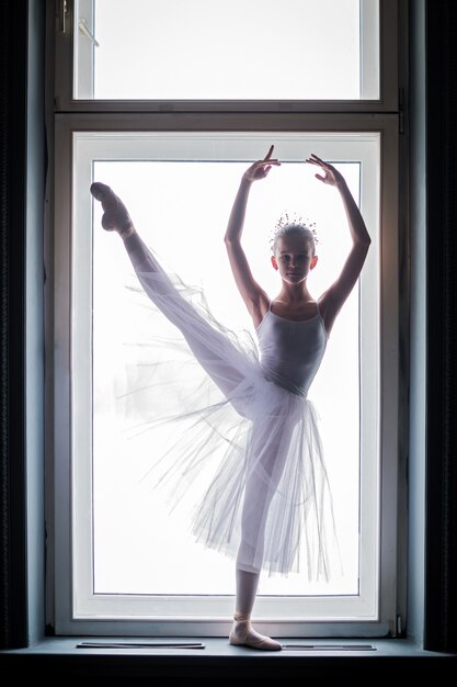 Young slender ballerina in a white tutu dances on pointe shoes in a spacious, bright room with large windows.