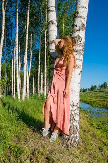 Young slender beautiful girl in a long coral dress posing in a birch grove smiling holding hand near the head