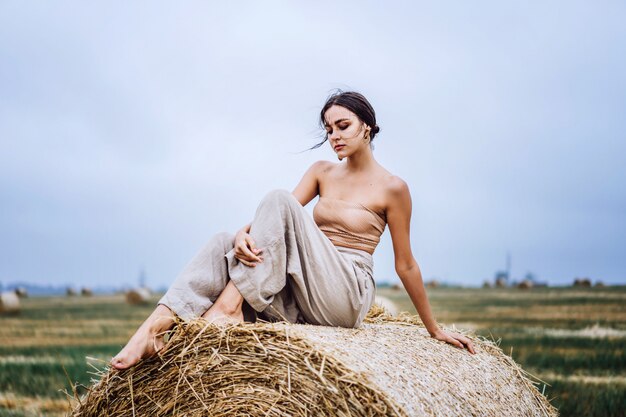 Young slender brunette girl, sitting on hay, posing for camera, photo shoot on field, blue sky.