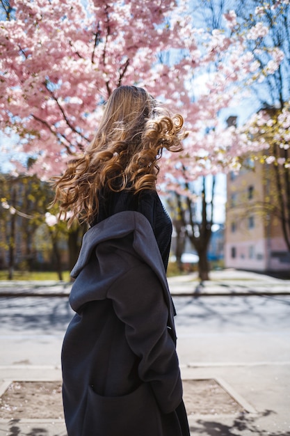 Young slender female model with long wavy hair and, dressed in a gray coat, sneakers, spinning on the street. Spring blossom trees woman girl laughs and runs to rejoice