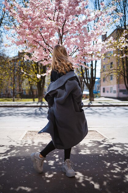 긴 물결 모양의 머리와 회색 코트, 운동화를 입고 거리에서 회전하는 젊은 날씬한 여성 모델. 봄 꽃 나무 여자 소녀 웃 고 기뻐서 실행