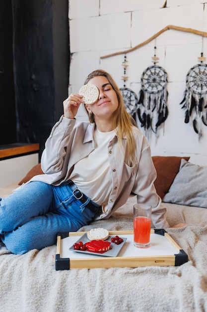 A young slender woman is sitting on the bed and eating a delicious breakfast of rice cakes and fresh juice Healthy Eating