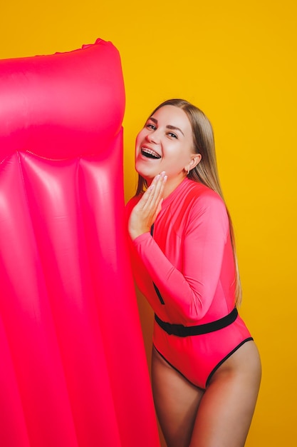 Young slender woman in a pink swimsuit on a yellow background Girl with an inflatable mattress for swimming in the pool Summer vacation