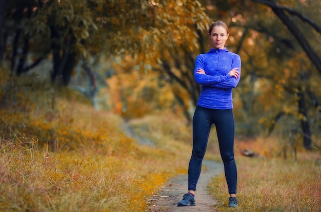 A young slim athletic woman trainer with crossed arms in a black leggins and blue jacket stands on a path in the autumnal forest.