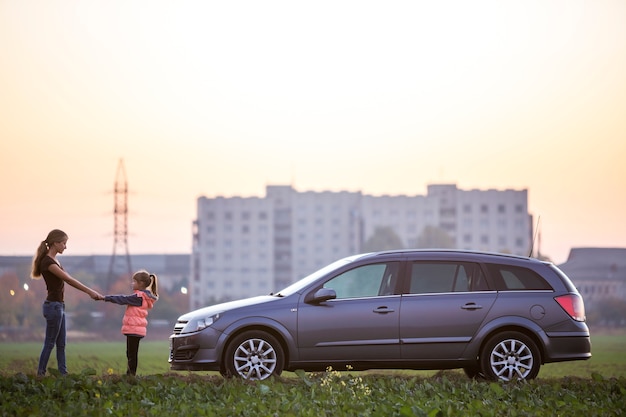 young slim attractive long-haired woman mother and small child girl daughter holding hands in green field at silver car on blurred apartment building and clear sky copy space background.