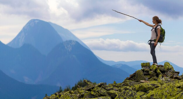 Young slim blond tourist girl with backpack points with stick at foggy mountain range panorama standing on rocky top on bright blue morning sky. Tourism, traveling and climbing concept.