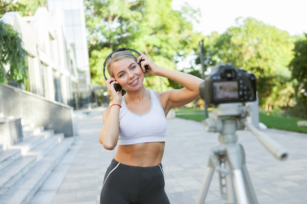 Young slim fit woman in sportswear with headphones filming fitness blog on camera outdoors