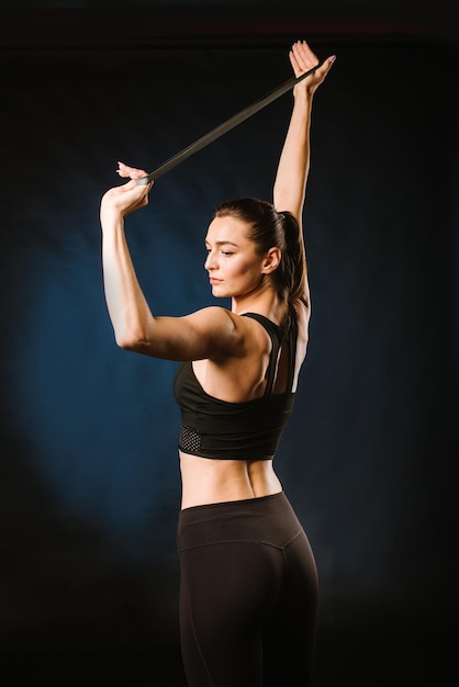 Young, slim girl doing exercises with rubber expander on black background.