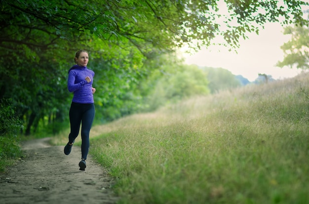 A young slim runner  in a black sports leggins and and blue jacket  fast runs along the path  on the  beautiful green forest. Photo show  active healthy lifestyle.