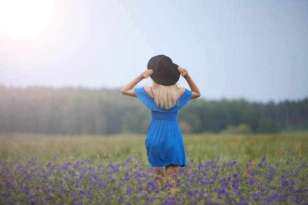 A young slim woman in a blue dress with a black hat walks in a flower field in summer