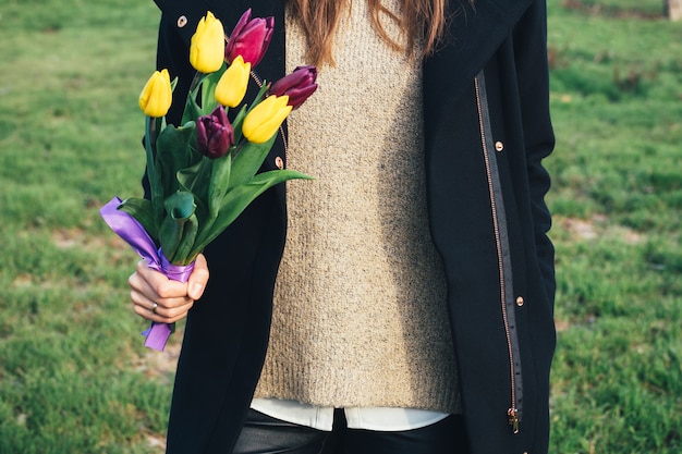 緑の芝生の上に立って、手にチューリップの花束を持ってコートの若いスリムな女性