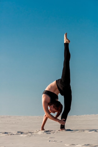 Young slim woman do gymnastic exercise at white sand beach under blue sky.