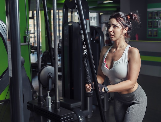 Young slim woman practicing extension of arms with ropes in exercise machine at gym