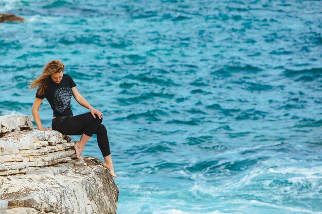 Young slim woman sitting on the cliff edge looking at sea copy space