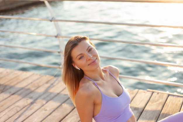 Young slim woman in sportswear enjoying music with earphones in her ears while sitting on beah pier. Sunrise Morning Workout Concept