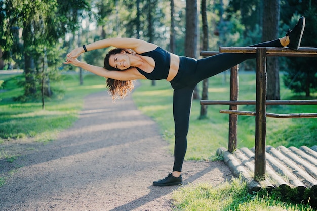 Young slim woman stretches outdoors demonstrates her flexibility wears cropped top and leggings leans on left poses outdoor in park makes aerobic exercises Active lifestyle and fitness concept
