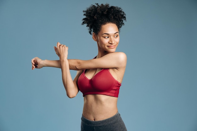 Young slim woman stretching her arms while standing isolated over blue background at studio and looking away