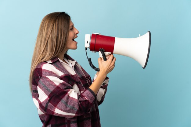 Young Slovak woman isolated on blue wall shouting through a megaphone to announce something in lateral position