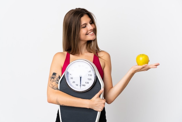 Young slovak woman isolated on white holding a weighing machine while looking an apple