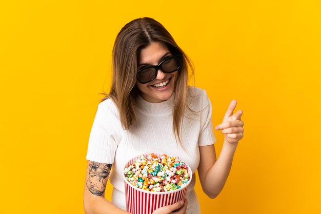 Young slovak woman isolated on yellow  with 3d glasses and holding a big bucket of popcorns while pointing front