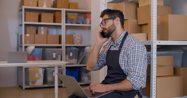 Young small business owner using laptop talking on phone in warehouse