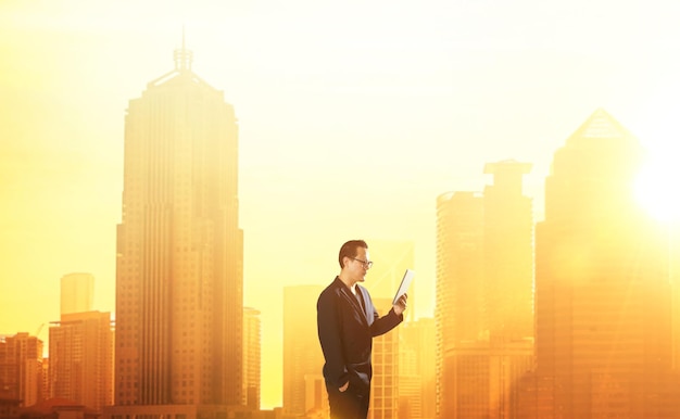 Молодой умный бизнесмен держит и использует цифровой планшет с размытым горизонтом города утром, стоя перед захватывающим горизонтом с переполненными корпоративными небоскребами
