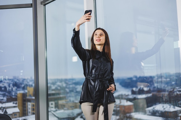 Young smart businesswoman in casual clothes working with phone while standing near window and reporting and writing notes and against blurred modern office interior