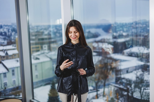 Молодая умная деловая женщина в повседневной одежде, работающая с телефоном, стоя у окна, репортаж и запись заметок на фоне размытого современного офисного интерьера
