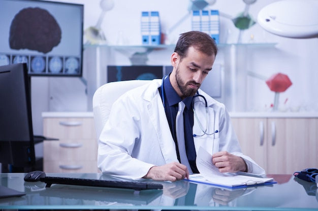 Medico giovane e intelligente che controlla la sua lista di pazienti sui suoi appunti. medico bello con cappotto bianco e stetoscopio.