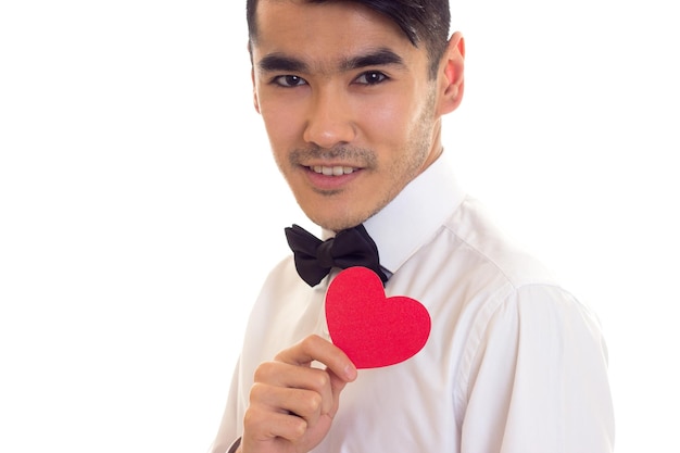 Молодой умный мужчина с черными волосами в белой футболке с черным галстуком-бабочкой держит красное бумажное сердце на белом фоне в студии