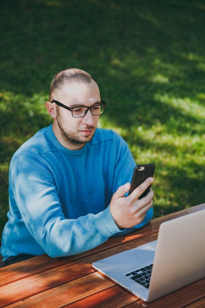 Giovane uomo d'affari o studente intelligente in camicia blu casual, occhiali seduti al tavolo inviando sms sul telefono cellulare o leggendo notizie nel parco cittadino utilizzando il computer portatile, lavorando all'aperto. concetto di ufficio mobile.