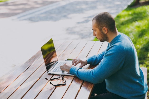 젊은 똑똑한 남자 사업가 또는 캐주얼한 파란색 셔츠 안경을 쓴 학생은 녹색 배경에서 야외에서 작업하는 노트북을 사용하여 도시 공원에서 휴대전화와 함께 테이블에 앉아 있습니다. 모바일 오피스 개념입니다. 측면보기.