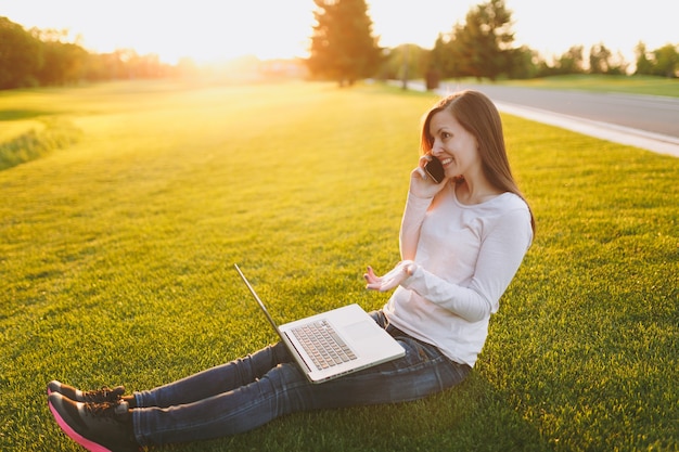 Young smart student female in casual clothes. Woman talking on mobile phone, working on laptop pc computer in city park on green grass sunshine lawn outdoors. Mobile Office. Freelance business concept